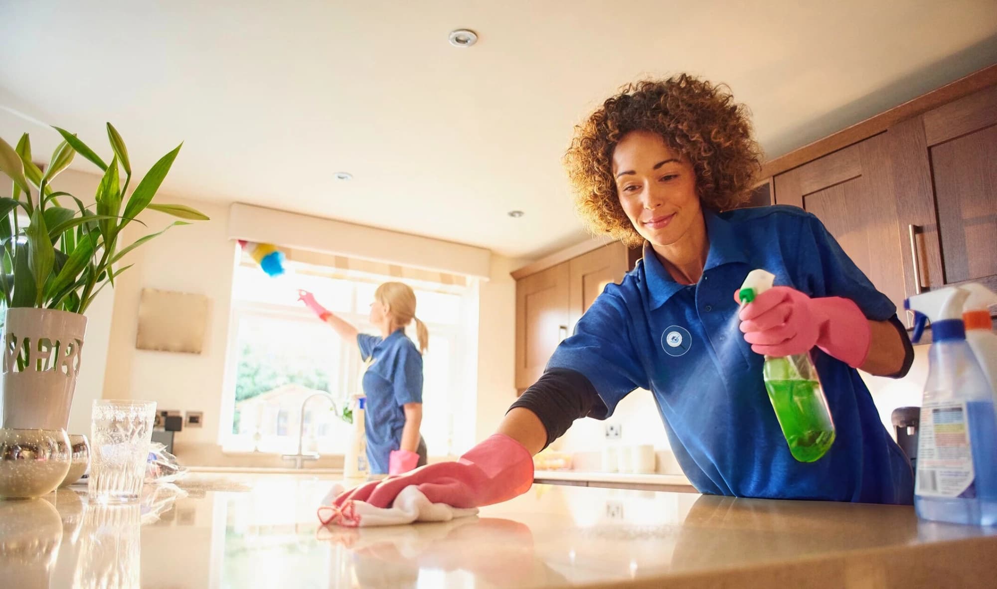 Two Magnified Cleaning cleaners in blue uniforms wiping a kitchen bench and window